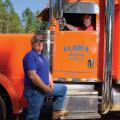 A man in a blue shirt stands in front of an orange semi-truck with another man in the driver seat.