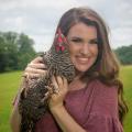 A young woman with brown hair and a pink shirt stands smiling while holding a black and white speckled chicken next to her face. 