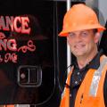 A man wearing a bright orange construction vest and hard hat stands in front of a logging machine.