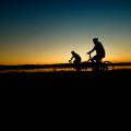 The silhouettes of two cyclists are seen in front of a blue and yellow sunrise that reflects on a lake below.