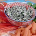 Spinach dip in a ceramic bowl centered on a plate surrounded by cut celery, baby carrots, and sliced red bell pepper.