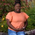 A smiling young girl wearing an orange shirt sits on a ledge in front of flowers.