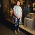 A smiling young girl wearing a floral blouse standing in front of a military museum exhibit.