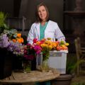 A woman wearing a white coat stands behind floral arrangements and a bucket full of flowers.