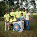 A man standing behind an archery target and surrounded by six children, all smiling.