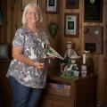 A woman, smiling, holding a 4-H flag and 4-H trophy and standing in front of several 4-H plaques affixed to the wall.