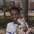 A young smiling Black man holding a snake and standing near a city street.