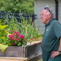 A man, standing beside a wooden table filled with plants, looking into the distance.
