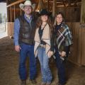 A teenage girl wearing a cowboy hat stand between her mom and dad, also wearing a cowboy hat, in a stable.