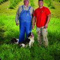 •	A man in overalls stands beside a man in a red T-shirt in front of a tree farm