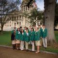 A group of high school girls and boys wearing khaki bottoms and green 4-H blazers stand in a group in front of the state capitol building.