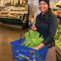A woman in a grocery store holding a blue basket filled with produce like leafy green lettuce.