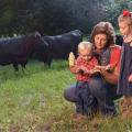 A woman and two small children in a field with black cows grazing behind them.