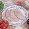 An array of ingredients in glass bowls, including romaine lettuce, onion, diced red pepper, Mexicorn, diced tomato, black beans, salsa, cooked diced chicken breast, and mozzarella cheese, surround a blue and white plate bearing four halves of whole wheat pita bread.