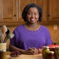 MSU Extension Agent Natasha Haynes, an African American woman with chin-length straight black hair stands behind a kitchen table displaying a crock of kitchen utensils, a red pot, and assorted home-canned and fresh fruits and vegetables. 