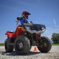 A teenage boy in a blue and white long-sleeved shirt and protective gear, including a red and black helmet and black gloves, rides a red ATV around orange cones during a safety class. (Kevin Hudson)