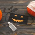 A black jack-o-lantern mask, bottle of sanitizer, and two pumpkins on a wooden backdrop.