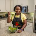 A woman in a kitchen smiles and holds a forkful of salad up for the camera.