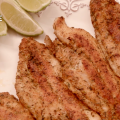Four browned, seasoned catfish fillets on a white plate garnished with lime wedges.