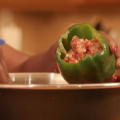 Hands spoon a ground meat and riced cauliflower mixture into a raw green bell pepper held over a stainless steel bowl. 