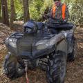 A hunter in camouflage and an orange vest places his rifle into storage on the back of an ATV in the woods.