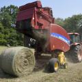 A red baler hitched to the back of an orange tractor drops a new, round bale of hay into a field.