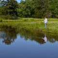 Older man holds a fishing pole on the shoreline while watching a red cork on a quiet pond.