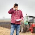 A farmer stands with a tractor in the background looking at a document and holding a hand to his head in worry.