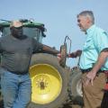 Two men facing each other in conversation and standing beside a tractor and equipment with a clear, blue sky overhead.