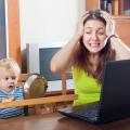A young mother grabs her head in frustration while working from home on a laptop computer as her baby looks on. 