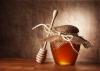 Wooden honey dipper lying against a small jar of honey that is covered with a decorative piece of fabric tied with a string.