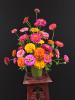 A arrangement of pink, orange and yellow zinnias sit on a stool against a black background.