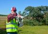 Man wearing a reflective safety vest looks at a white drone he is holding at shoulder height. A toppled pine tree and empty agricultural field are in the background.