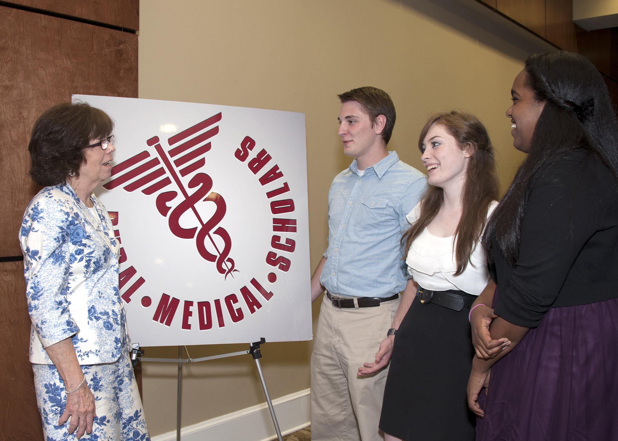 An instructor and three students have a discussion in front of a Rural Medical Scholars sign.
