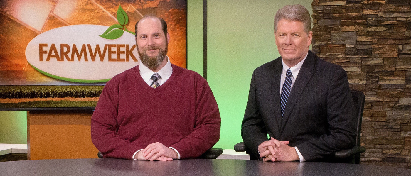 Two men sit at a table with a TV in the background that reads 'Farmweek'