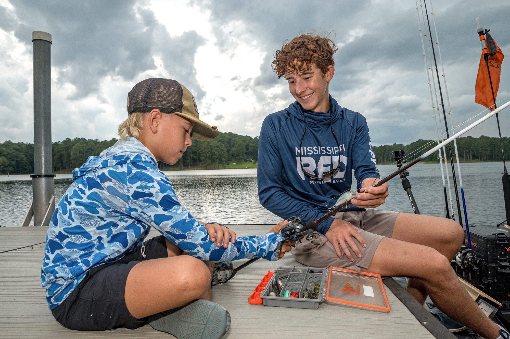 A young child at a lake holding a fishing rod while an older child supports it.