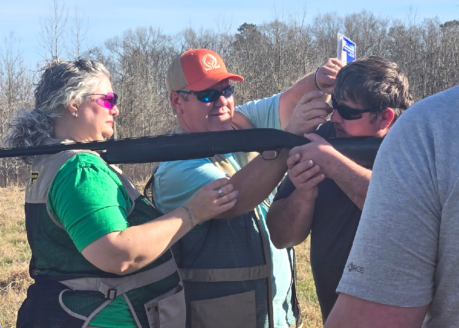A person rests a gun on a person’s shoulder while she and another person look on.