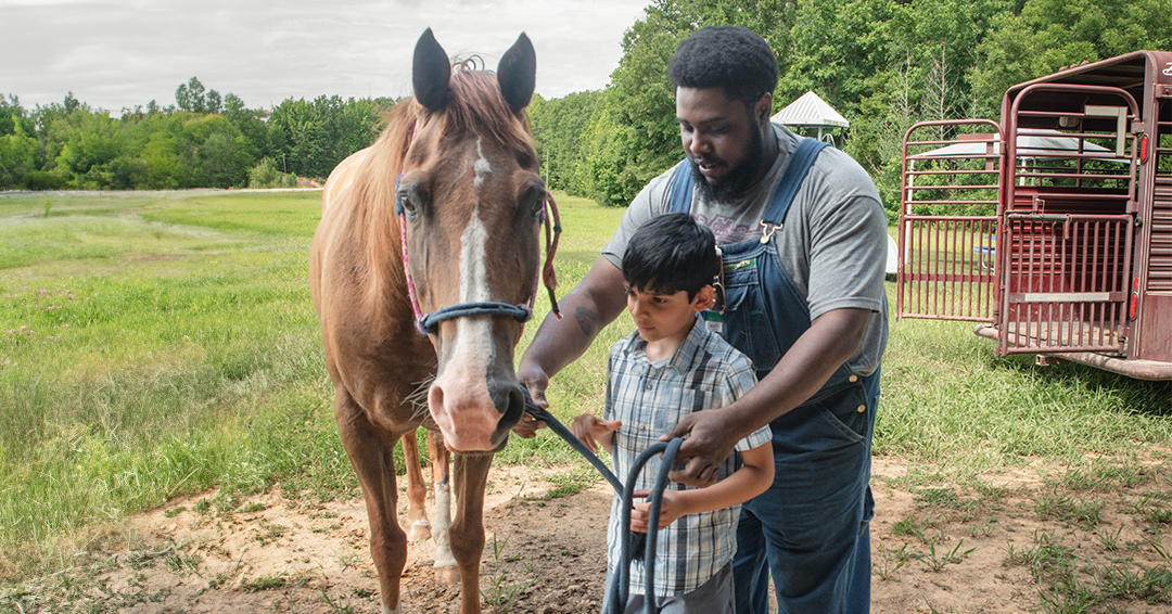 A man helps a boy with tacking for a horse.