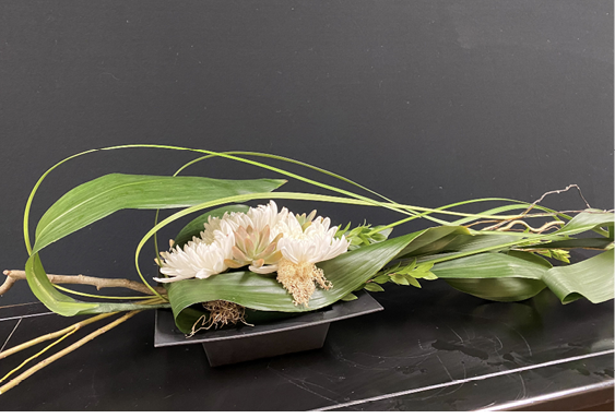 A floral arrangement, featuring a mix of flowers, glossy Aspidistra leaves, arching liriope, and corkscrew willow, is laid out on a counter.