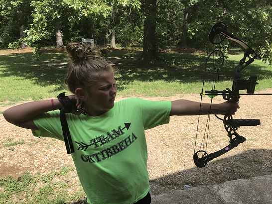 Young archer holds the bow straight toward the target with the bow arm extended and the drawing hand on the string so the body forms a T shape.