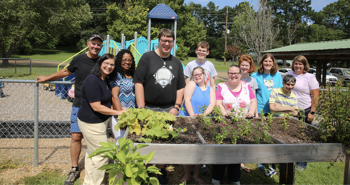 Adults and children stand behind a raised garden bed outside.