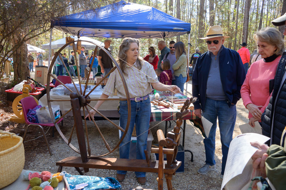 A person holds thread from a spinning wheel as other people watch.