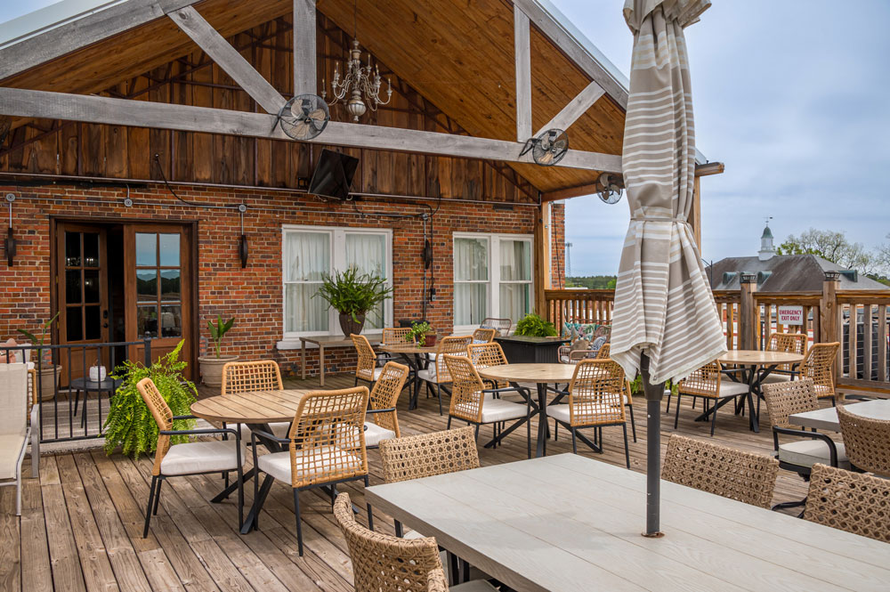 White tables with wicker chairs on a deck outside a brick building.