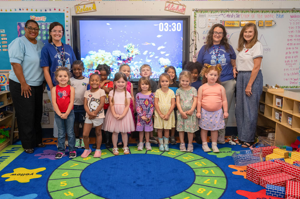 A group of children standing in a classroom and four adults standing behind them, two on each side.