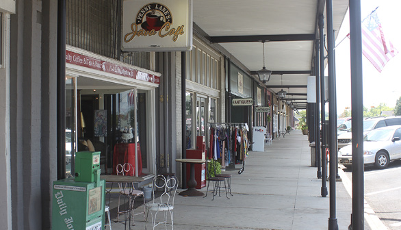 Covered walkway featuring storefront signs and displays, including tables and chairs, newspaper dispensers, and a rack of clothes.