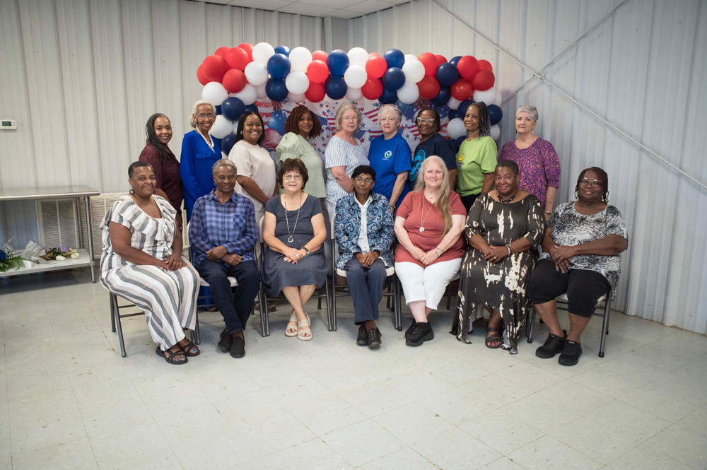 A group of smiling people standing and seated in front of a red, white, and blue backdrop.