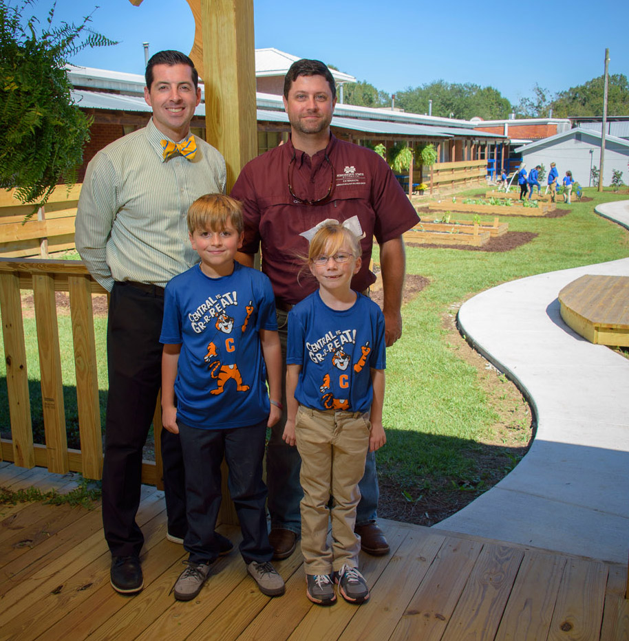 Two men, one on the left in a white &amp; blue striped polo and one on the right with a maroon polo, stand behind a male and female child, both blonde and wearing T-shirts with “Central is Gr-r-reat!” and a tiger printed on them. They all stand on a wooden patio in front of a small farm site.