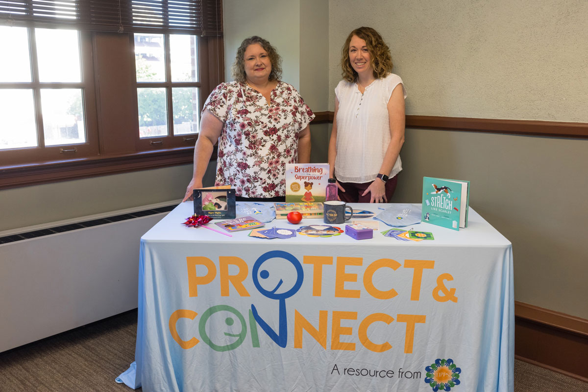 Two women stand behind a table with a cloth that reads “Protect &amp; Connect.”