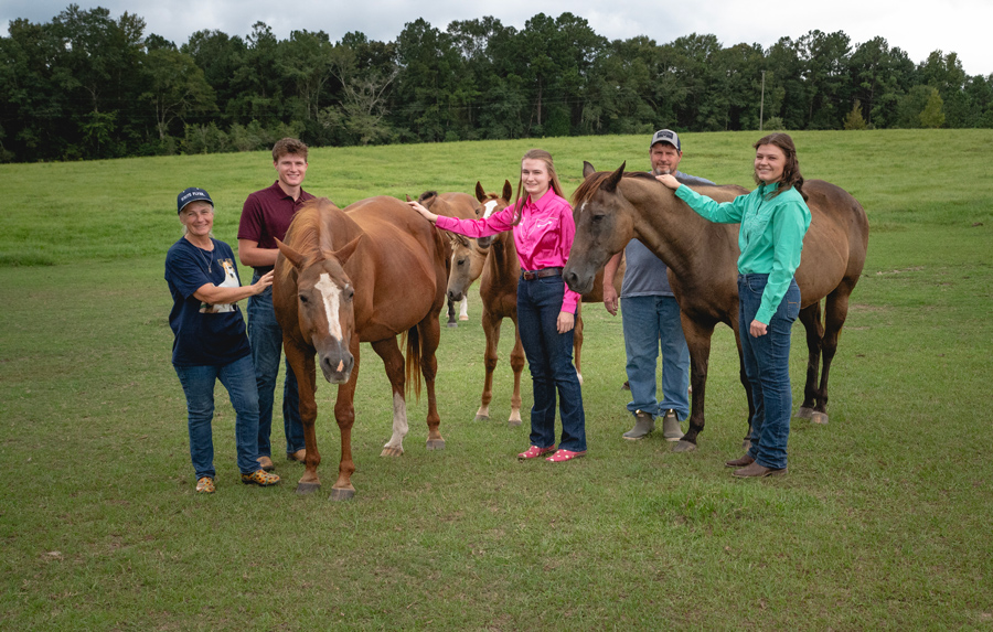 Five people standing with four horses in a field.
