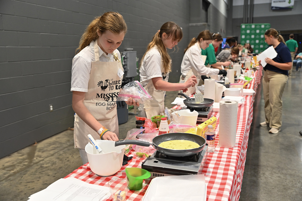 A student holds an ingredient bag at their cooking station while other students stir a pan of eggs at their stations. 